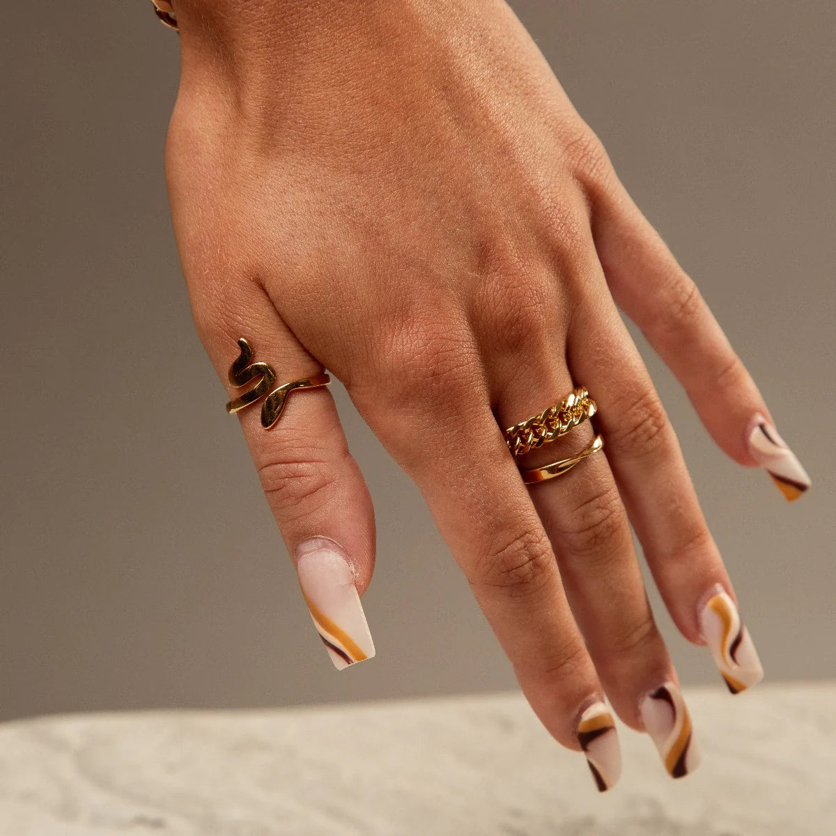 Hand wearing gold tone Bruges Ring and other gold rings with stylish patterned nails on a neutral background